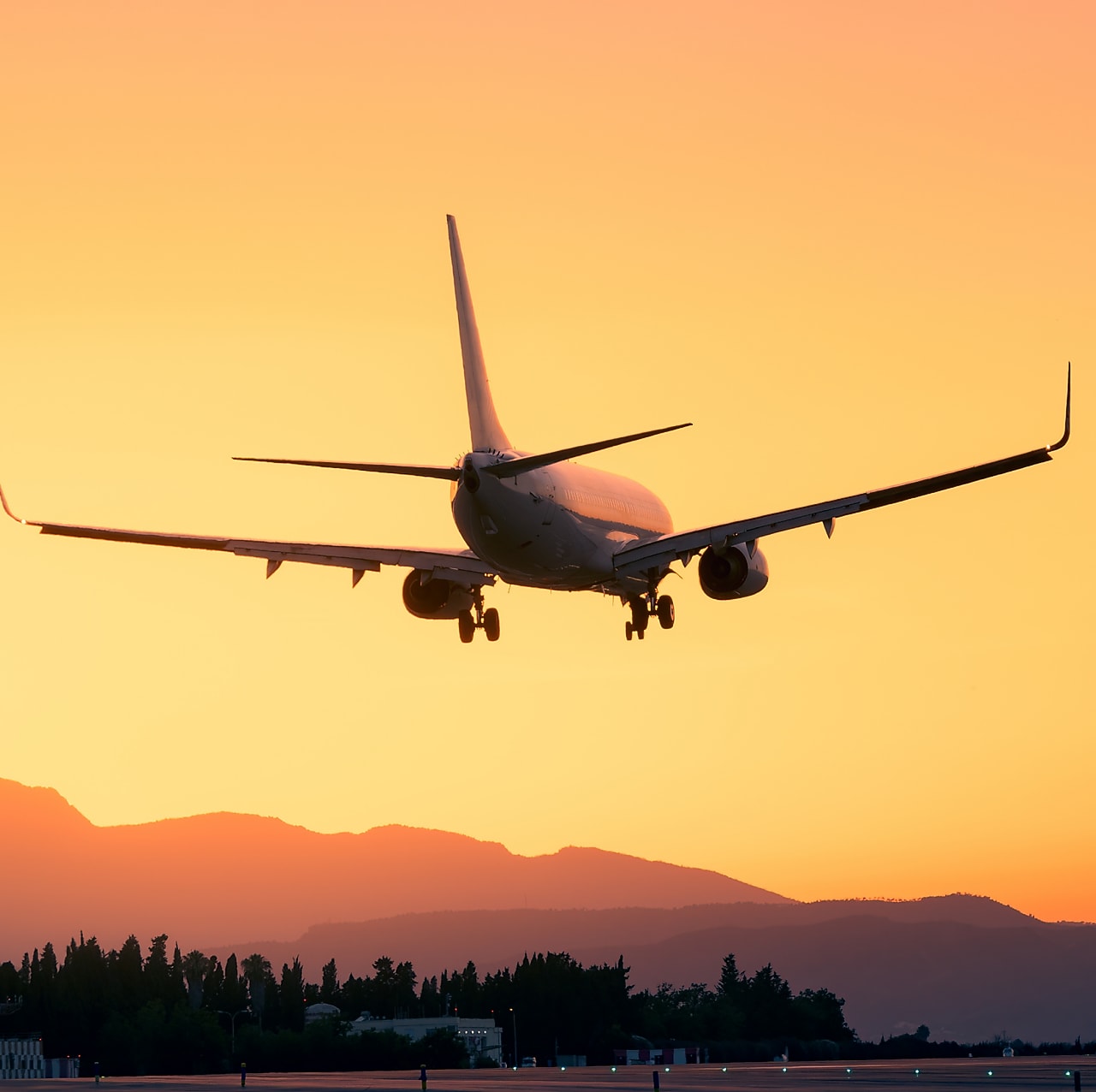 A commercial airplane coming in for landing against a vibrant orange sunset sky, with silhouettes of mountains and trees in the background.