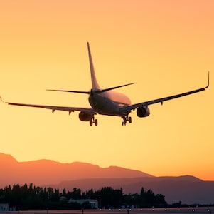 A commercial airplane coming in for landing against a vibrant orange sunset sky, with silhouettes of mountains and trees in the background.