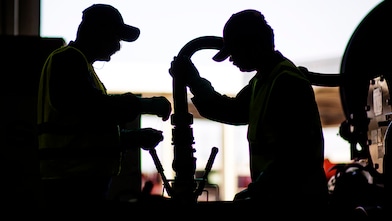 Silhouetted view of two workers in high-visibility vests operating fueling equipment.