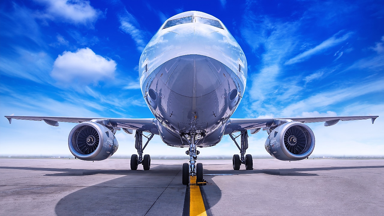 Front view of a commercial airplane parked on an airport runway under a bright blue sky with scattered clouds.