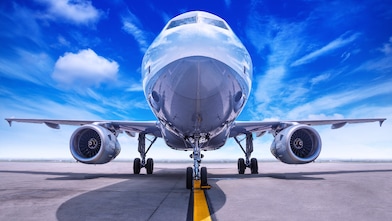 Front view of a commercial airplane parked on an airport runway under a bright blue sky with scattered clouds.