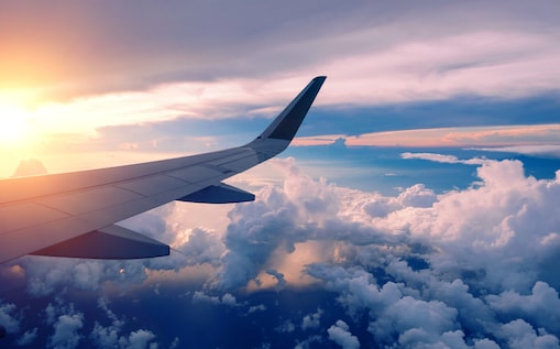 View from an airplane window showing the wing above a sea of clouds during a vibrant sunset, with warm sunlight illuminating the sky and reflecting off the cloud tops.
