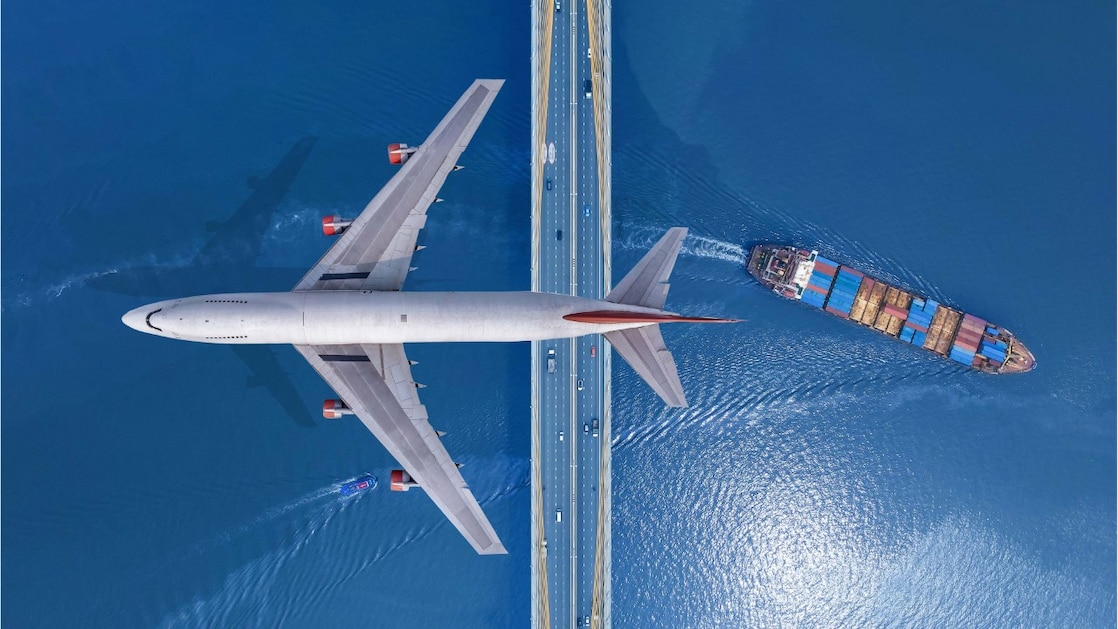 Aeriel shot of a plane flying over a bridge with a cargo ship passing under the bridge to the right