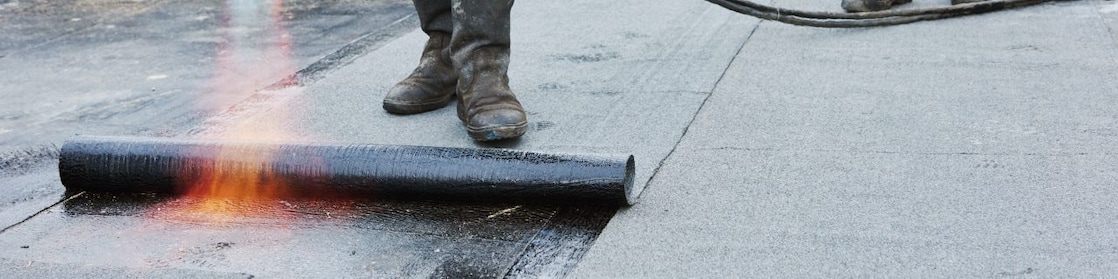 Flat roof installation. Heating and melting bitumen roofing felt by flame torch at construction site