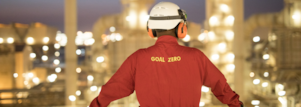 A worker wearing a hardhat and red shirt with “Goal Zero” embroidered on the back standing in front of a blurred refinery at night.