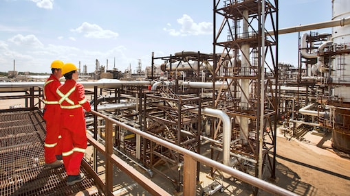 Two workers in red jumpsuits and yellow hardhats look over refinery in the background