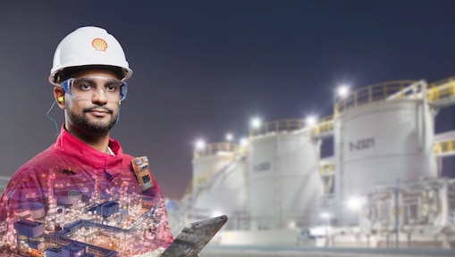 Bearded worker in hard hat, safety glasses, and a red Shell jumpsuit in front of a plant at night.