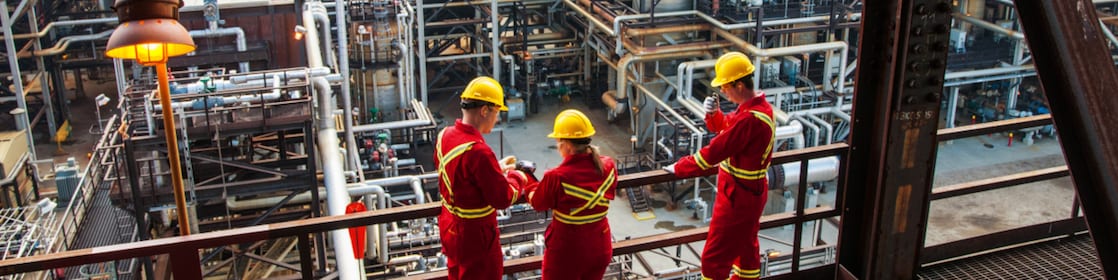 Three workers in red jumpsuits and yellow hardhats discussing sulphur recovery while standing on a platform looking over a refinery.