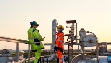 Two workers in yellow and orange safety gear discussing the Claus Off-Gas Treating (SCOT) process while standing on refinery against a bright, grey sky