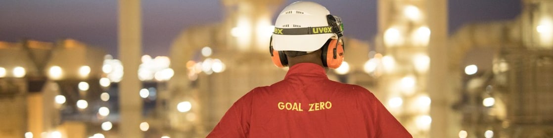 A worker wearing a hardhat and red shirt with “Goal Zero” embroidered on the back standing in front of a blurred refinery at night.