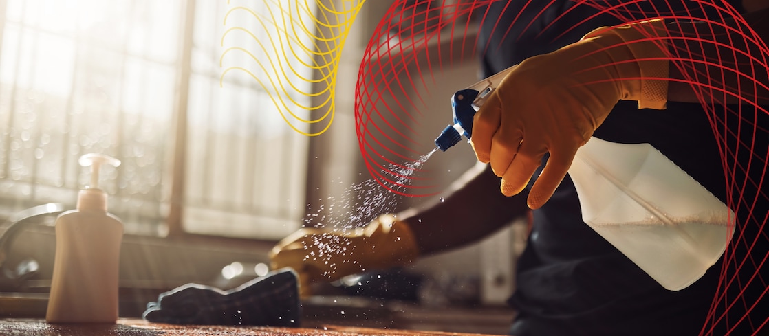 Gloved hand spraying cleaning solution onto a surface in a sunlit room.