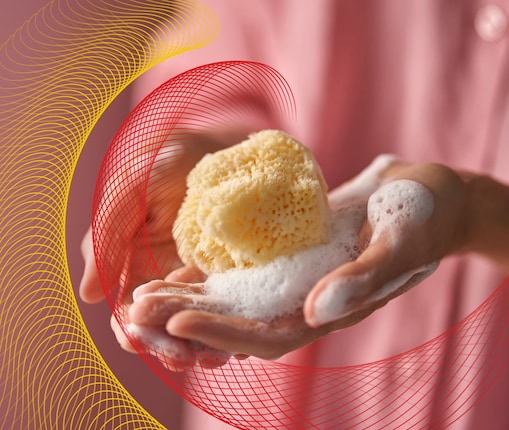 Hand holding a natural sponge covered in soap suds against a soft pink background.