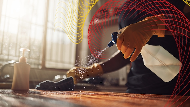 Gloved hand spraying cleaning solution onto a surface in a sunlit room.