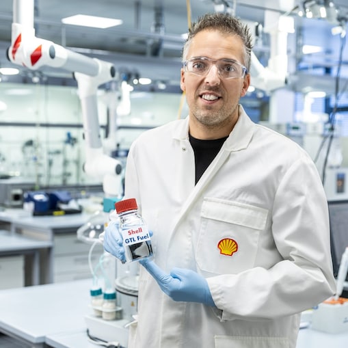 a person in a white coat and goggles holding a bottle of Shell GTL fuel
