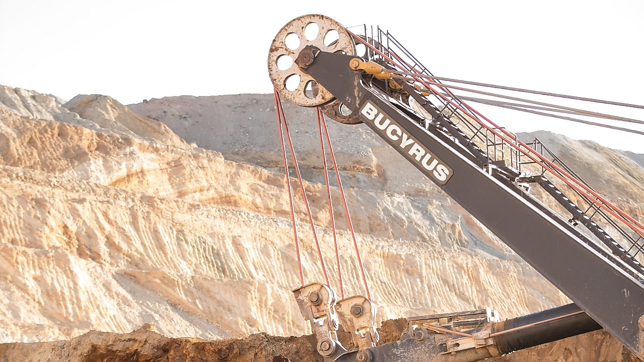A large mining excavator arm with cables and pulleys operating in an open-pit mine, with layered rock formations visible in the background.