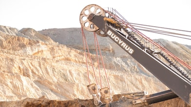 A large mining excavator arm with cables and pulleys operating in an open-pit mine, with layered rock formations visible in the background.