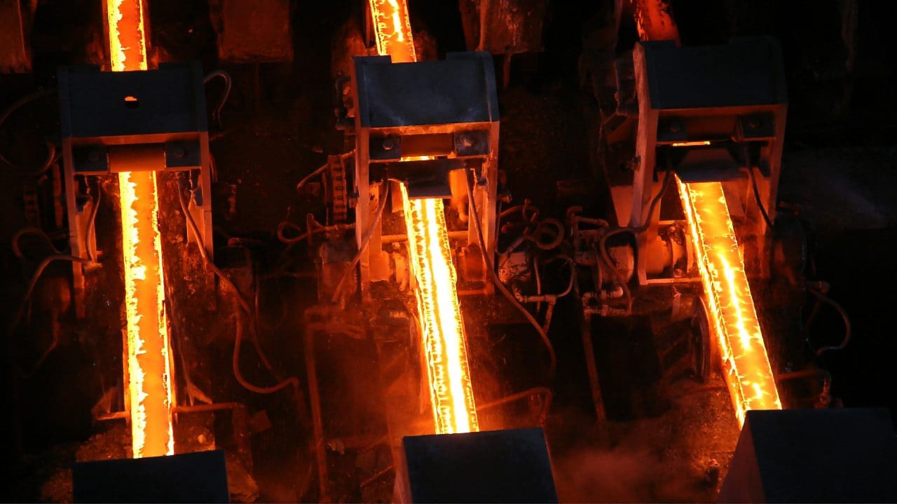 Metal, glowing orange under heat, is being processed into long slabs in an industrial steel manufacturing facility.