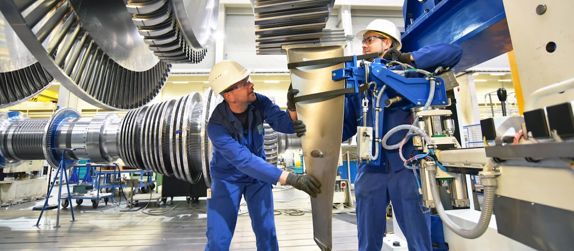 Two engineers on a wind farm, one wearing a yellow hard had and the other in a white hard hat. Both engineers are looking at a tablet.