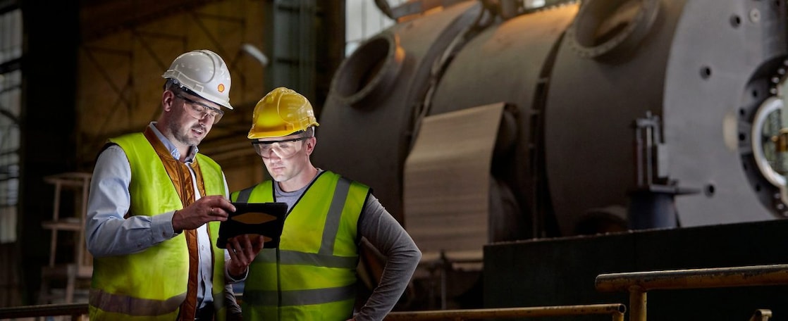 2 men in high vis jackets, hard hats and safety glasses in foreground, large industrial machinery in background