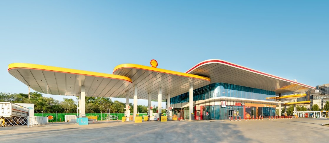 Modern Shell fuel station with a curved yellow and red canopy, forecourt pumps, and an attached convenience store under a clear blue sky.