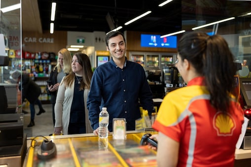 Man smiling at a Shell Select store counter with a bottle of water and snack on the counter, a Shell staff member serving him and two women browsing in the background.