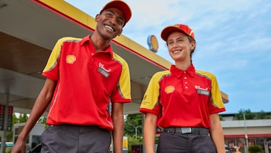 Two smiling Shell forecourt staff in red and yellow uniforms standing outside a Shell station under a partly cloudy sky.