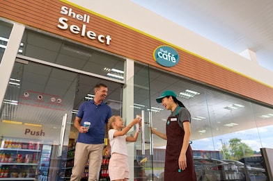 Father and daughter smiling outside a Shell Select store with a Shell Café sign, greeted by a smiling staff member in a green cap and apron holding the door open.