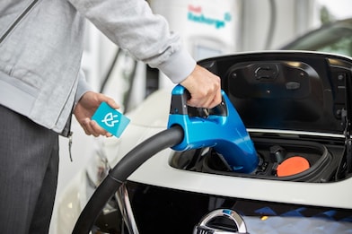 Close-up of a hand inserting a blue charging connector into an electric vehicle's charging port while holding a contactless charging card.