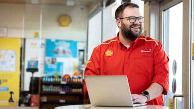 Shell forecourt employee in a red V-Power shirt smiling while typing on a laptop inside a Shell store.
