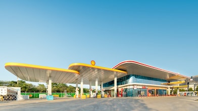 Modern Shell fuel station with a curved yellow and red canopy, forecourt pumps, and an attached convenience store under a clear blue sky.