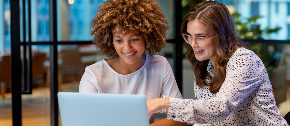 Two women in an office looking at a laptop screen together, one pointing at the display, with a coffee cup on the desk.