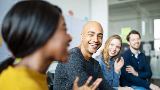 Group of colleagues laughing together during an informal office meeting, with sticky notes and a whiteboard in the background.