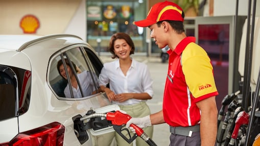 Shell forecourt attendant in a red and yellow uniform fuelling a white car while a smiling female customer stands nearby.