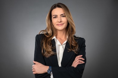 Woman with long brown hair in a dark blazer, arms crossed, smiling at the camera against a grey studio background.