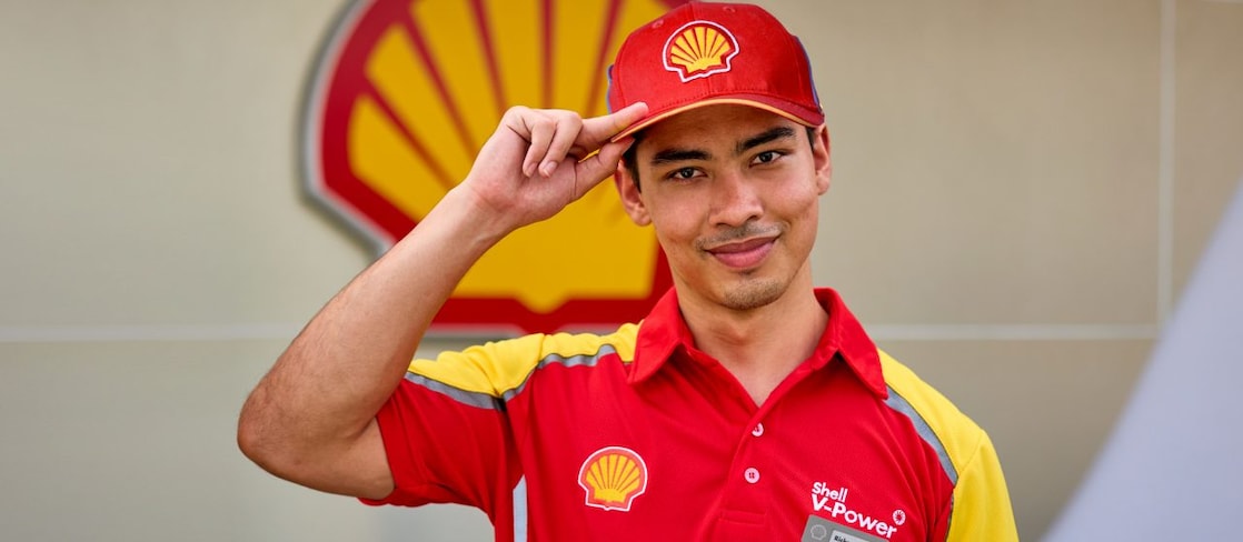 Young Shell forecourt employee named Ricky in a red V-Power polo shirt and cap, smiling and tipping his cap in front of a large Shell logo.