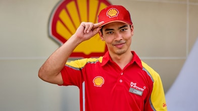 Young Shell forecourt employee named Ricky in a red V-Power polo shirt and cap, smiling and tipping his cap in front of a large Shell logo.
