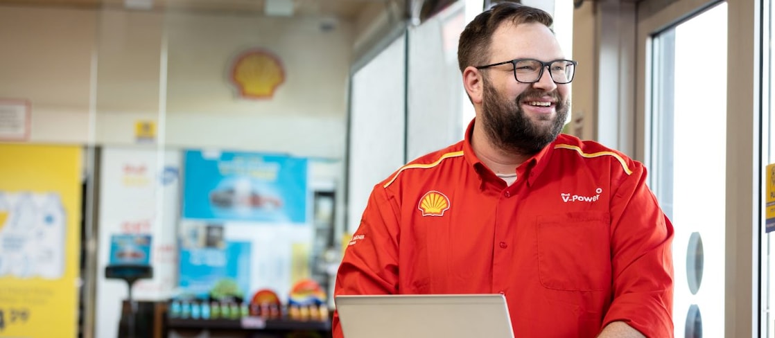 Shell forecourt employee in a red V-Power shirt smiling while typing on a laptop inside a Shell store.