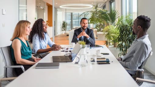 Four colleagues in a glass-walled meeting room having a discussion around a table with laptops, coffee cups and notebooks, with plants and city views in the background.