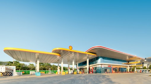 Modern Shell fuel station with a curved yellow and red canopy, forecourt pumps, and an attached convenience store under a clear blue sky.