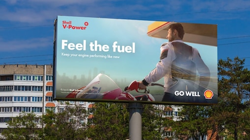 Shell V-Power billboard advertisement reading 'Feel the fuel, keep your engine performing like new', showing a motorcyclist refuelling at a Shell station, mounted above a row of residential buildings.