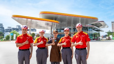 Five Shell staff in red, yellow, and brown branded uniforms giving a thumbs up together outside a Shell station with a curved canopy roof.