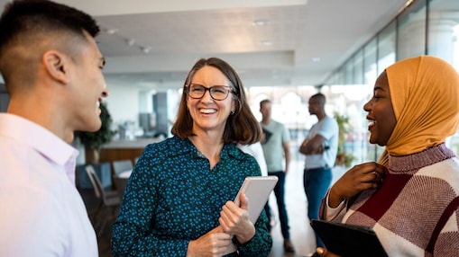 Three colleagues talking and smiling in an office setting, with one woman in a hijab and another holding a tablet, and blurred coworkers in the background.