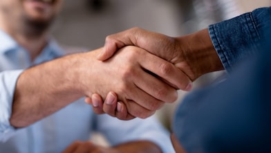 Close-up of two people shaking hands, one in a light blue shirt and one in denim, with a blurred smiling face in the background.