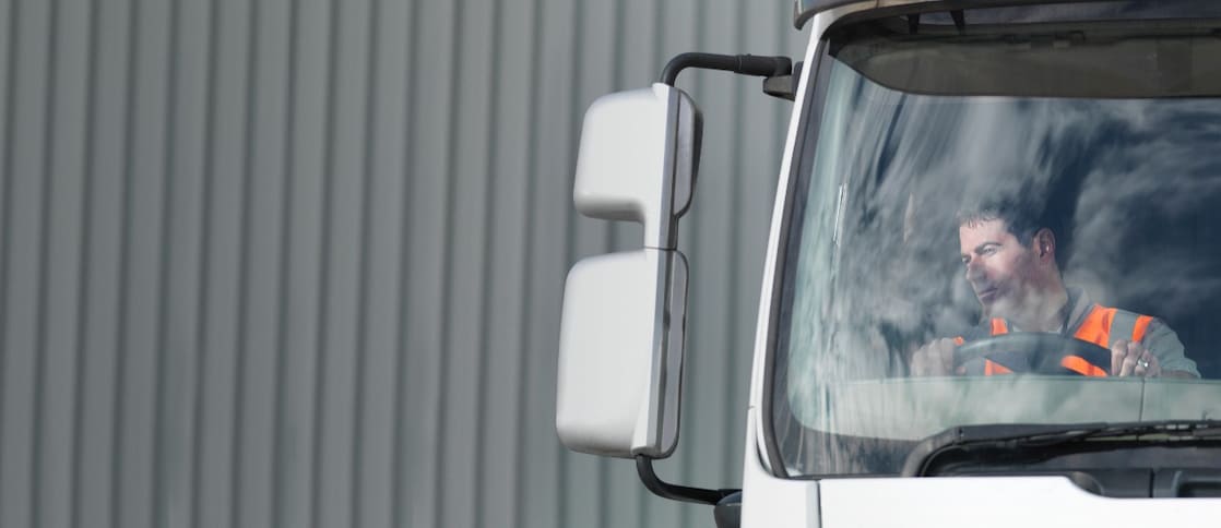 Driver sat at the steering wheel of a white truck looking out the right mirror wearing an orange high-vis jacket