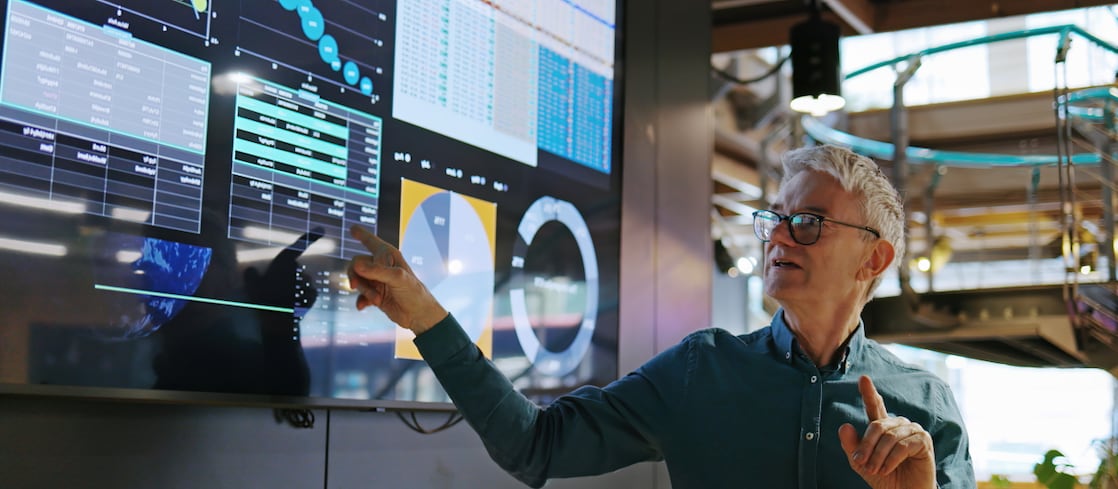 An engineer or data analyst presenting information on a large digital screen displaying charts, graphs, and data visualizations inside a modern industrial or research facility.