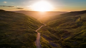 Winding road cutting through green hills at sunset, with the sun glowing over the horizon and casting warm light across the landscape.