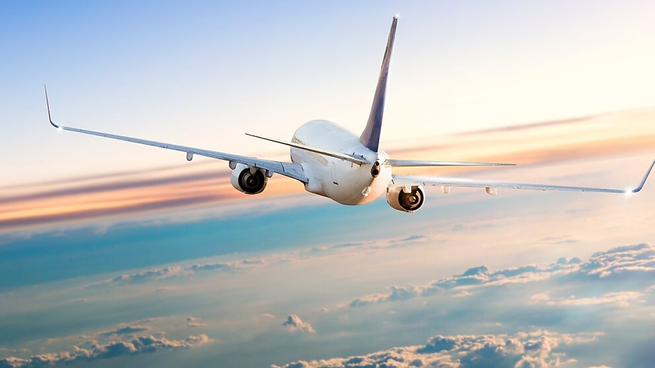 Rear view of a commercial airplane flying above clouds at sunset, with wings extended and a soft gradient sky in the background.