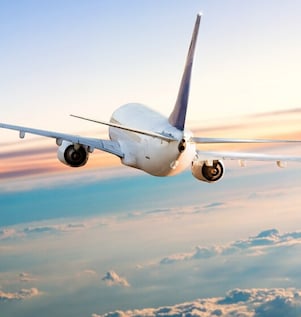 Rear view of a commercial airplane flying above clouds at sunset, with wings extended and a soft gradient sky in the background.