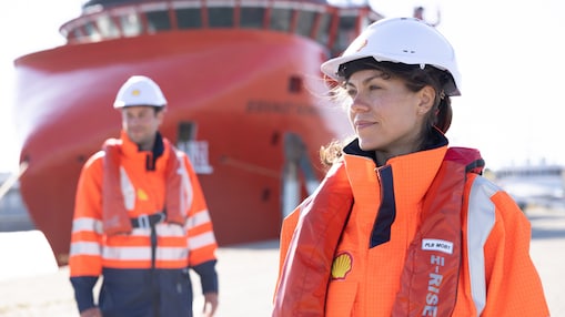 Two workers in safety helmets and high-visibility Shell uniforms with life vests, standing near a large red ship at a dock