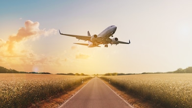 A commercial airplane flies low over a straight rural road surrounded by golden wheat fields at sunset. The sky glows with warm orange and yellow hues, with scattered clouds and soft sunlight illuminating the scene, creating a sense of motion and tranquility.